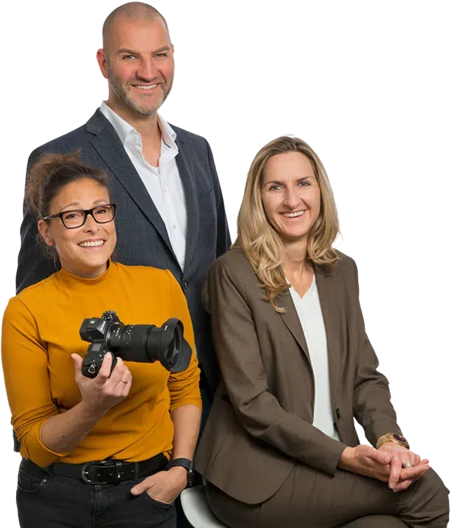 Drei lächelnde Geschäftspersonen posieren im Studio, eine Frau hält eine Kamera.